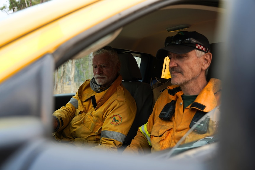 two men dressed in firefighting equipment sit in the front driver and passenger seats of a vehicle