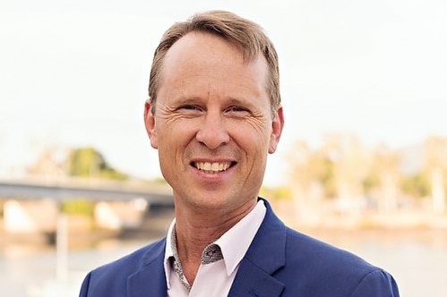 A man with brown hair smiles. He is wearing a navy suit jacket and a white shirt. There is a river and bridge in the background.