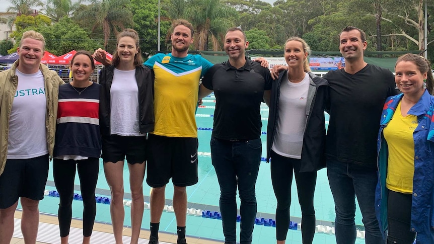 A group of eight athletes stand side-by-side with arms around each other as they stand beside a pool.