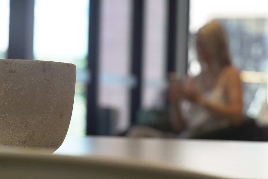 A woman with blonde hair is shown sitting on a chair out of focus in the background. In the foreground is a cup on a table.