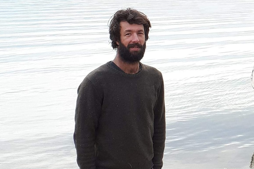 A man with a beard and dark hair stands on a shore with his hands in his shorts pockets.