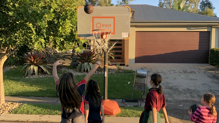 Four girls throw basketballs at a temporary hoop set up on the nature street in front of a house.
