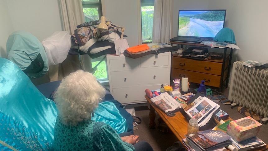 A de-identified grey-haired woman, shot from back, sitting on her couch in a room crammed with clothes, dresser, watches tv.