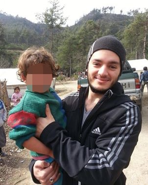 Man in black tracksuit and beanie smiles, holding young child, with trees and hill in background