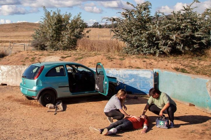 A person lying on red dirt near a small green car, being helped by two people.