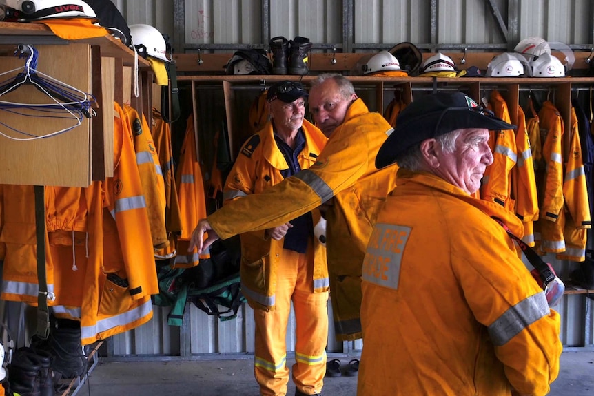 Three men wearing firefighting gear stand in a shed surrounded by more equipment.