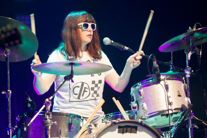 Tobi Vail drums at the 02 Academy Brixton in London. She is wearing a sunglasses and a white t-shirt with a grid on it