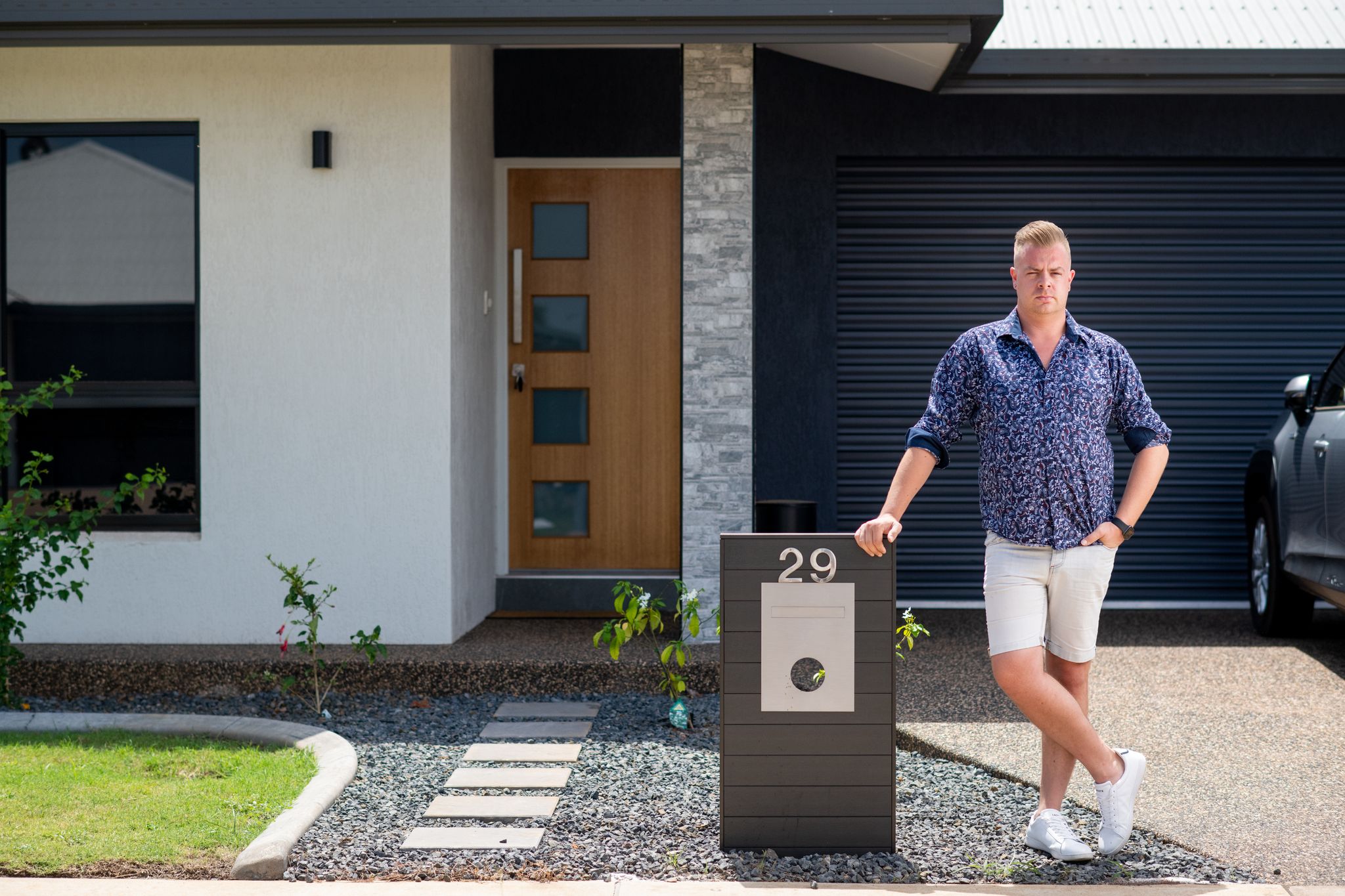 A blonde man wearing a navy collared shirt and white pants stands outside a property.