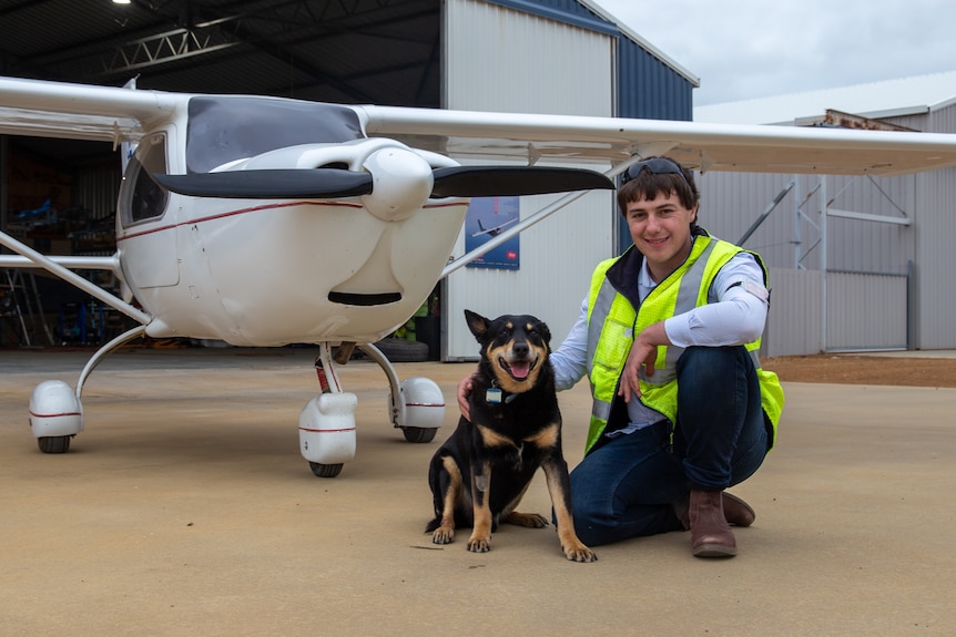A young man sits next to his dog in front of a light aircraft.