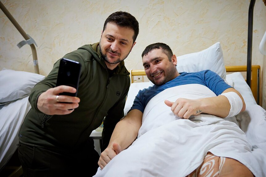 A man in khaki takes a selife with a wounded man in a hospital bed