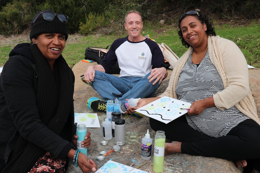 Two indigenous women, with man in the middle, all sitting on rock with paints and artwork 