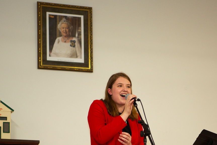 A young woman in a red jacket sings at the microphone in a small country hall.