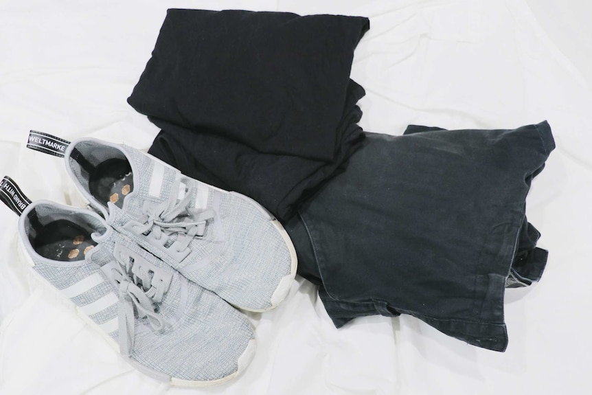A pair of grey sneakers sits next to a pair of black jeans and a pile of black shirts to depict a capsule wardrobe.