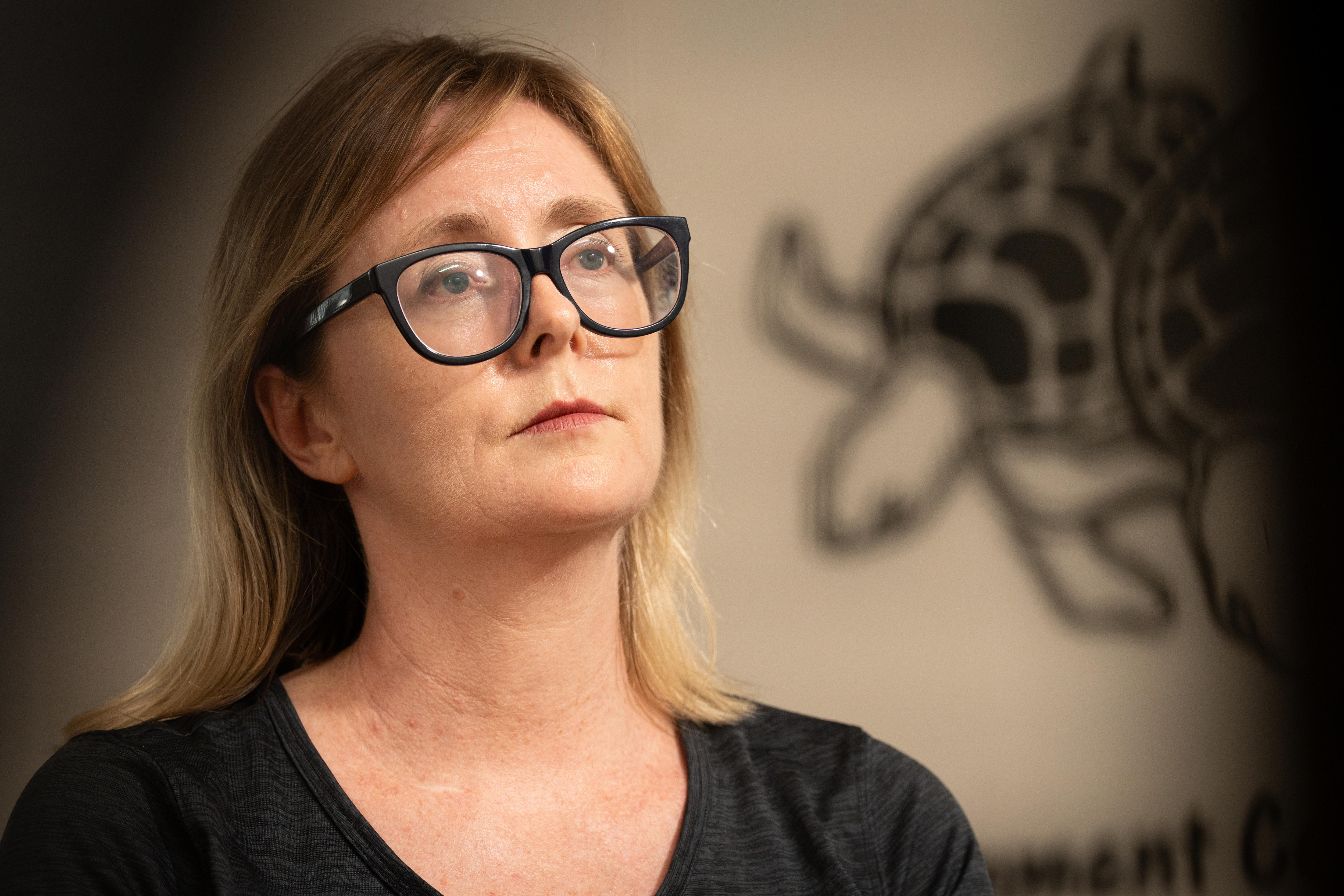 Un primer plano de una mujer blanca, cabello rubio/castaño fino hasta los hombros, blusa negra, ilustración de una tortuga en la pared detrás de ella.