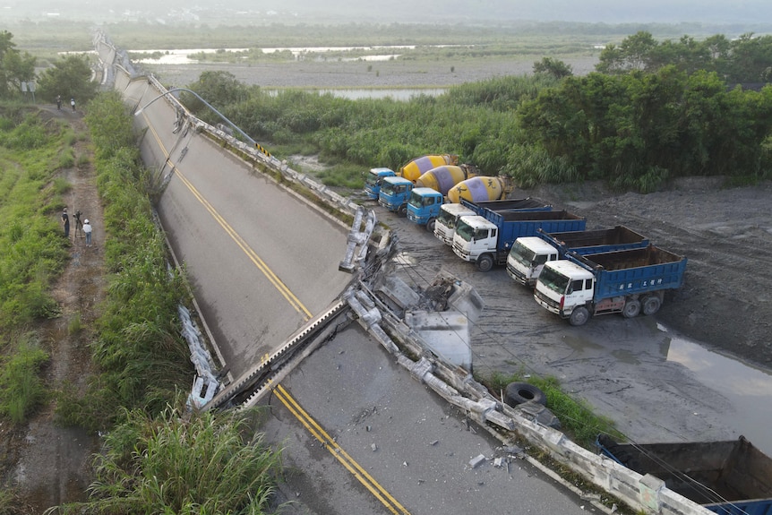 Multiple trucks parked on the side of the road next to a highway bridge that has fallen on its side. 