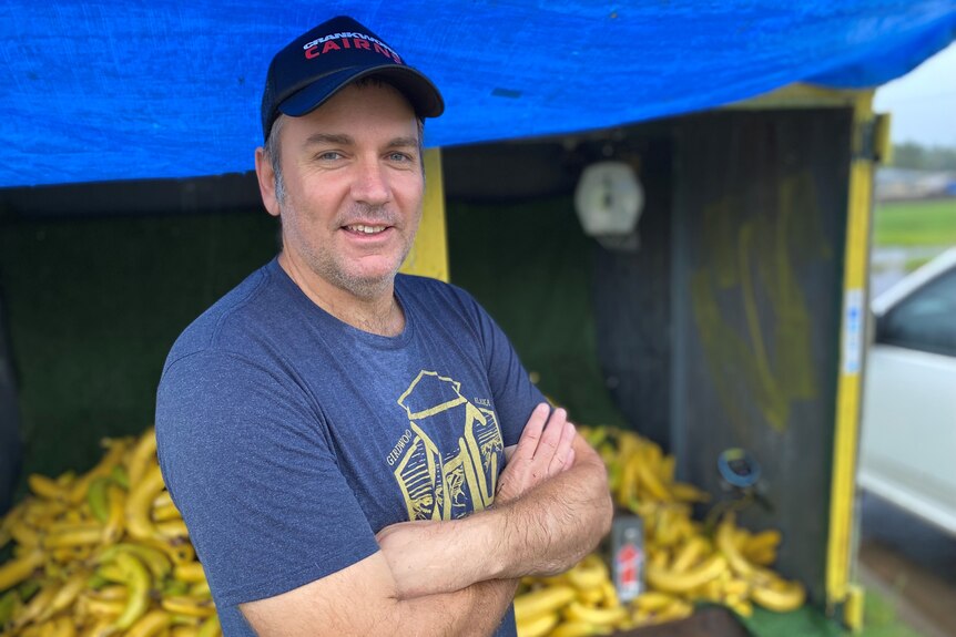A man wearing a cap stands in front of a trailer full of bananas.
