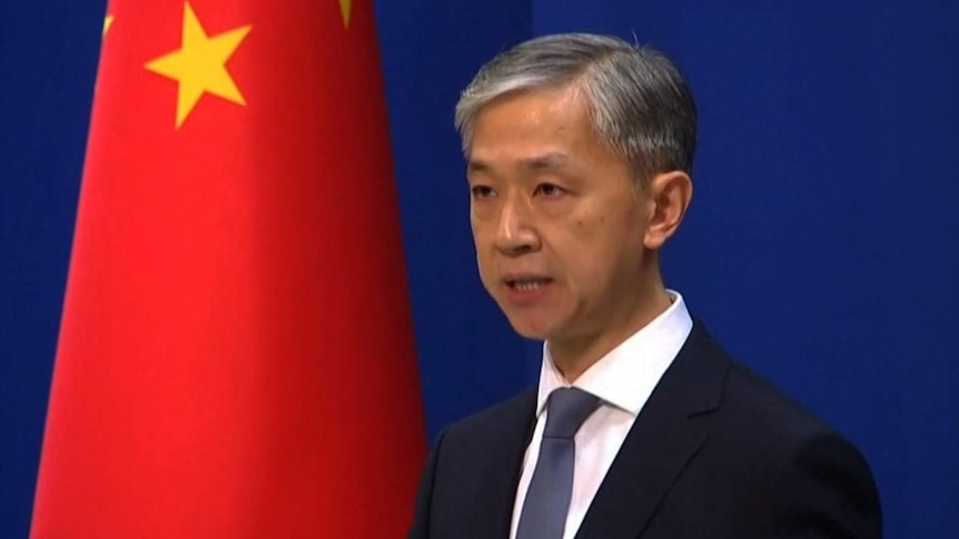 Man in suit and tie with grey hair stands in front of blue background and China flag