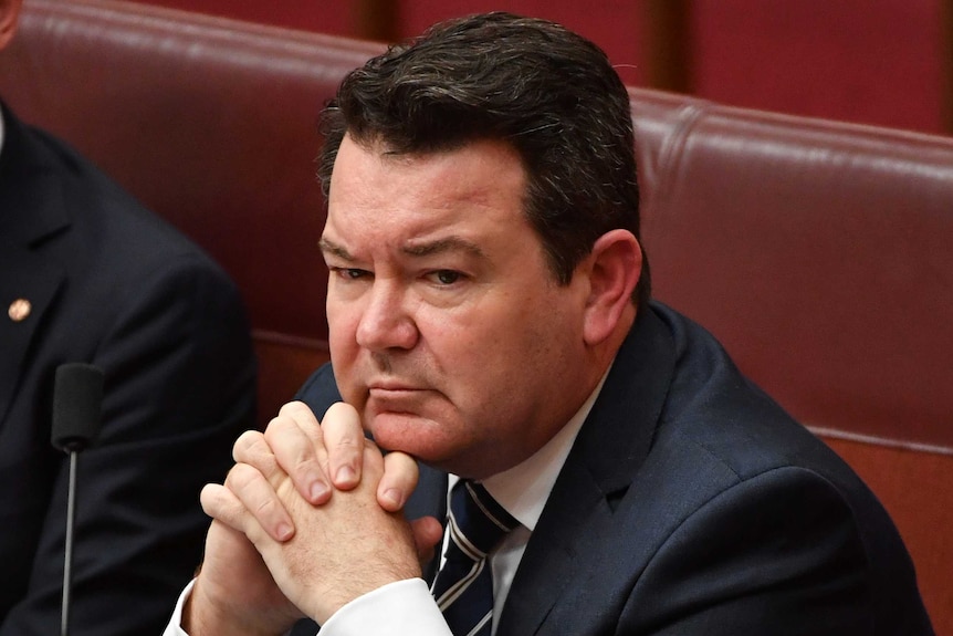 Liberal senator Dean Smith sitting in one of the red chairs of the Senate with his hands clasped under his chin