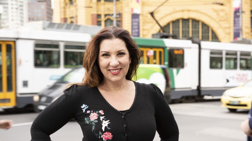 A smiling woman stands in front of Flinders Street Station, while a tram passes by.