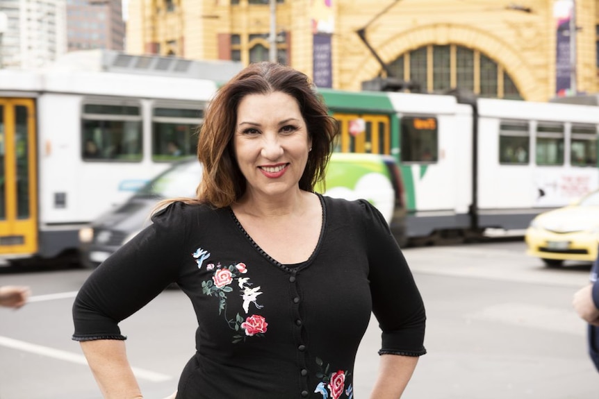 A smiling woman stands in front of Flinders Street Station, while a tram passes by.