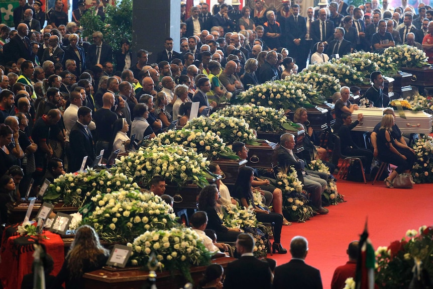 People watch the funeral services, standing amongst a row of 19 flower covered coffins.