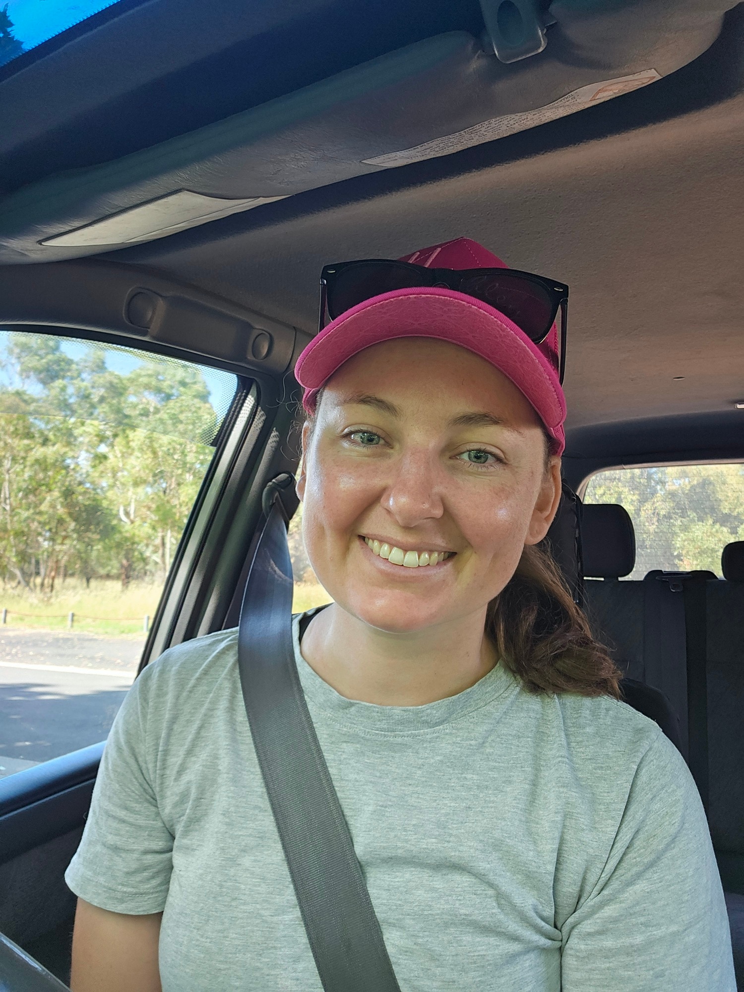 A smiling woman wearing a baseball cap sitting in a car with a seatbelt on.