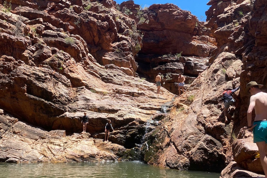 Swimmers climb or stand around the red rockfaces and a small waterfall cuts a path in the middle of the image. Hint of blue sky