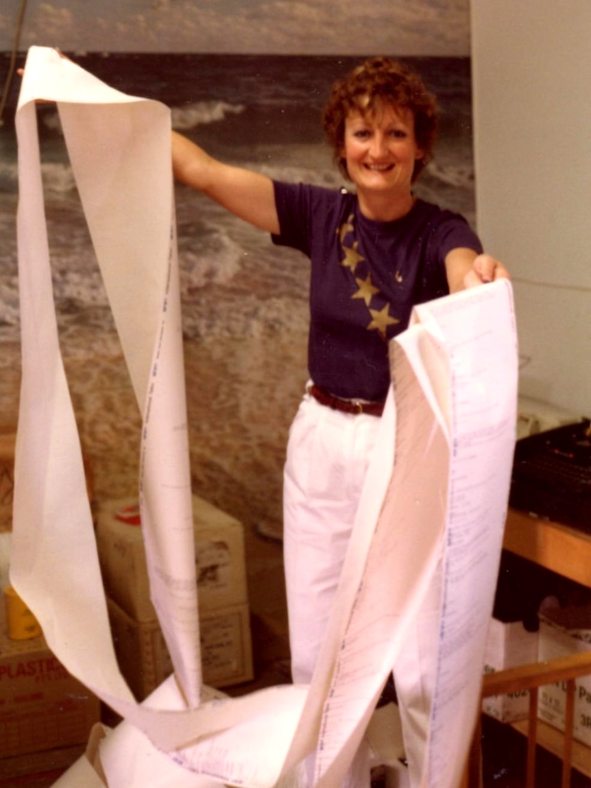 A smiling woman holds up reams of paper spewed forth from a telex machine.