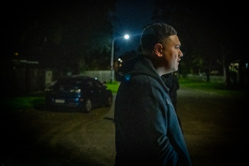 A young man walks on a suburban street at night. He wears a hoodie and is looking away from the camera.