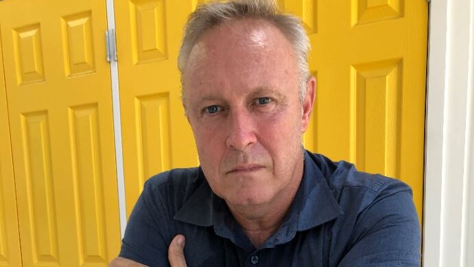A man sitting down with arms folded in front of a yellow door