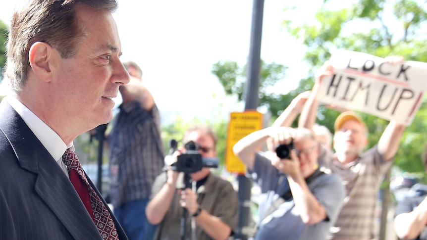 Paul Manafort, standing side on to the camera, walks past photographers and a man holding up a sign that says "lock him up".