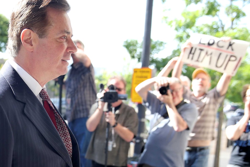 Paul Manafort, standing side on to the camera, walks past photographers and a man holding up a sign that says "lock him up".