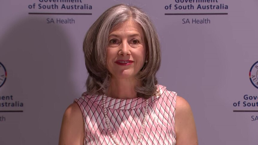 A woman wearing a sleeveless pink dress smiles in front of an SA Health banner