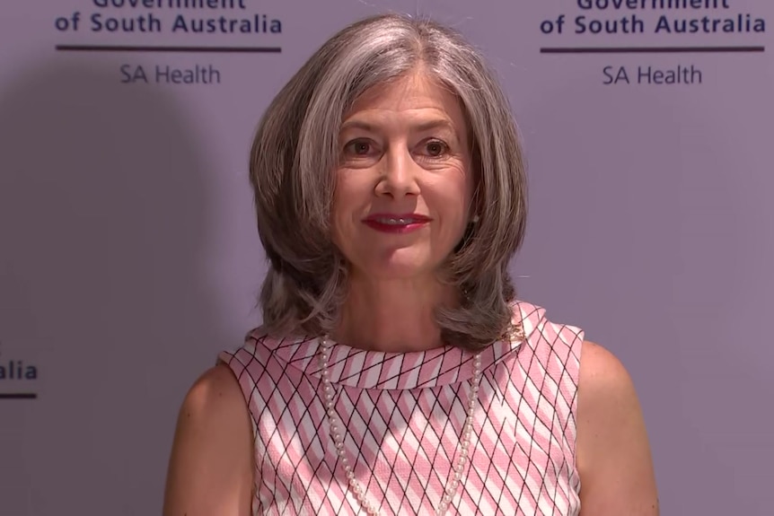 A woman wearing a sleeveless pink dress smiles in front of an SA Health banner