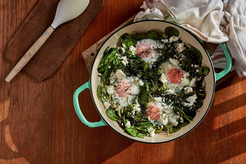 A wide pan with baked eggs sitting on top of baby spinach, leeks and broccolini, with feta and dill scattered on top, dinner.