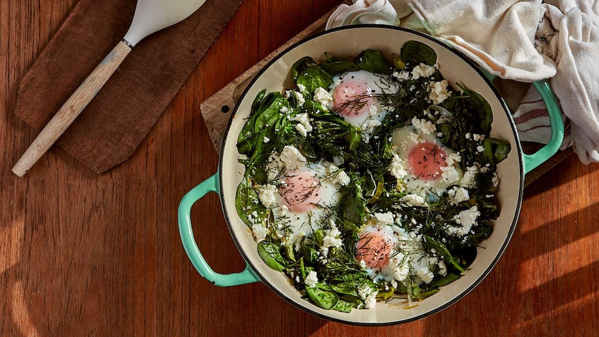 Pan with baked eggs sitting on top of baby spinach, leeks and broccolini, with feta and dill scattered on top, a dinner recipe.