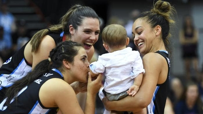 Three WNBL players celebrate with one holding a baby.