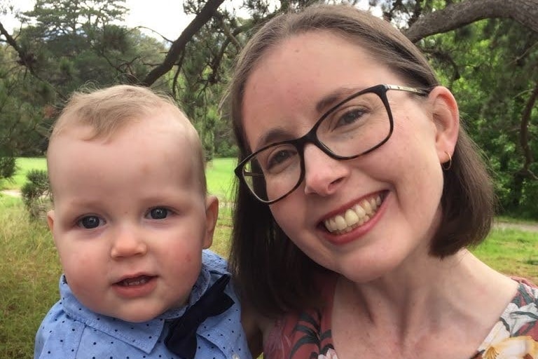 A brunette woman smiles as she holds up a blonde  baby