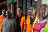 Five women wearing orange vests look straight ahead with concern on their faces.