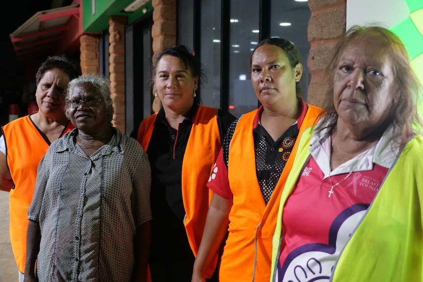 Five women wearing orange vests look straight ahead with concern on their faces.