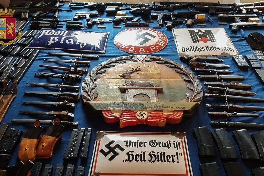 Guns and ammunition seized by Italy police surround a large Nazi plague with Swastika.