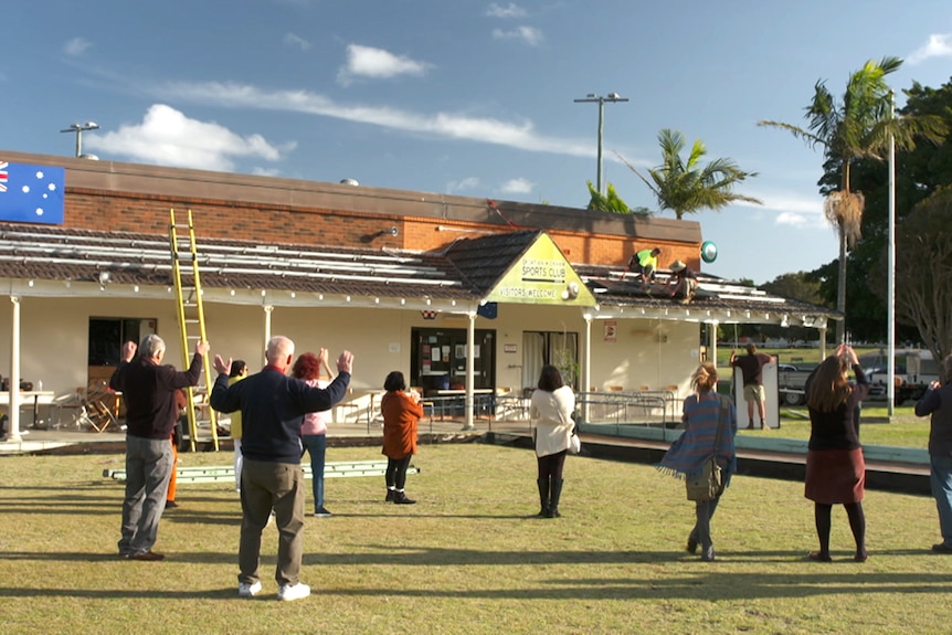 People on a bowling green look on as solar panels are installed on a roof