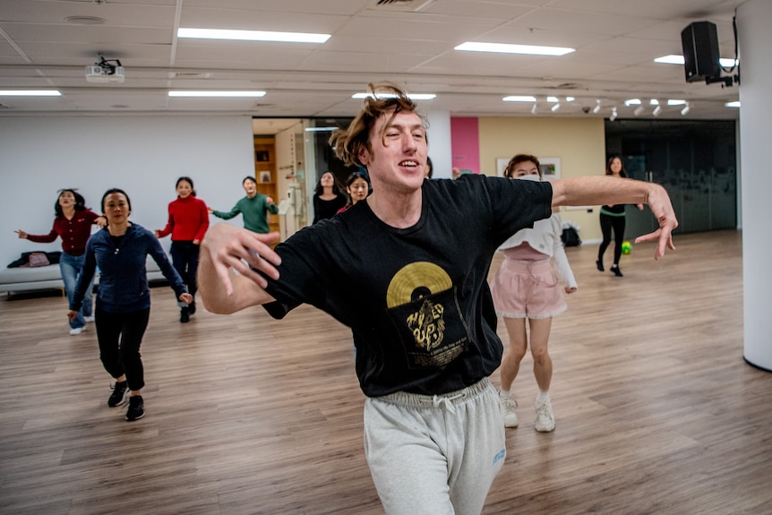 A person is teaching a dance class, with arms outstretched and people behind following.
