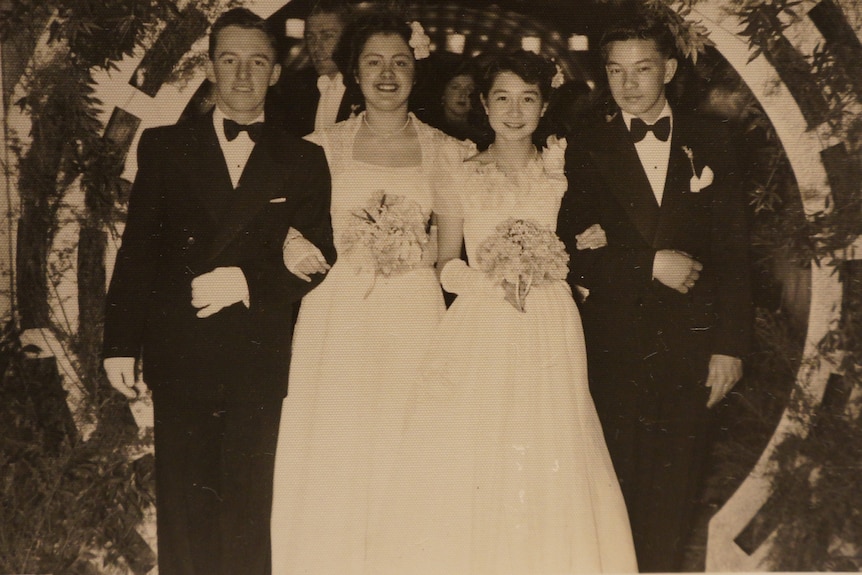 Two couples stand next to each other, both of the women in white dresses and the men in tuxedos