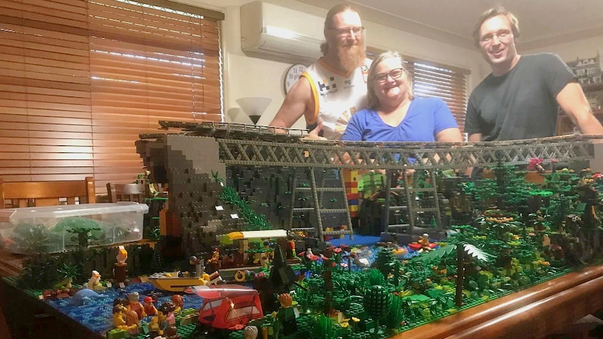 Three people standing  behind a large Lego display of a rail bridge surrounded by rainforest.