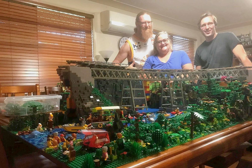 Three people standing  behind a large Lego display of a rail bridge surrounded by rainforest.