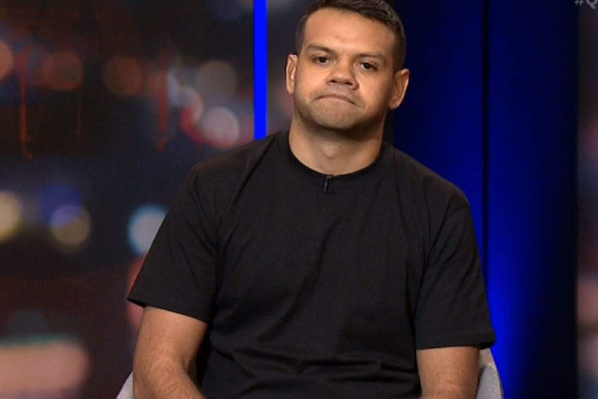 A man in a black t-shirt with black hair stares forward, frowning