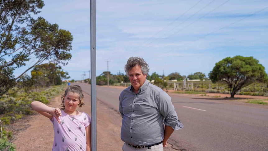 An 11-year-old girl with her thumb down with a middle-aged man next to a sign post with a missing label