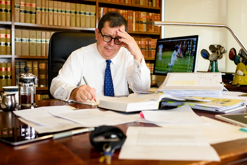 A man in a shirt and tie sitting at a big desk poring over books, with his head resting on his hand.