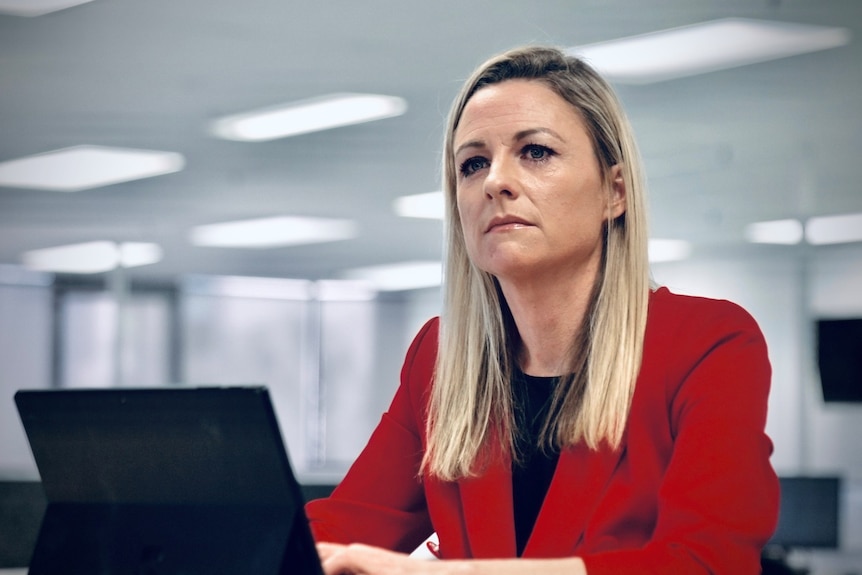 A woman with straight blonde hair wearing a red blazer and black top sits in an office in front of a black laptop.